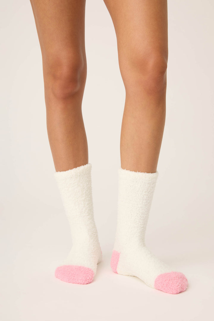 A person wears P.J. Salvage Doggone Cold Fuzzy Dog Crew Socks—white with pink toes and heels—standing on a light-colored surface, with only their lower legs and feet visible.