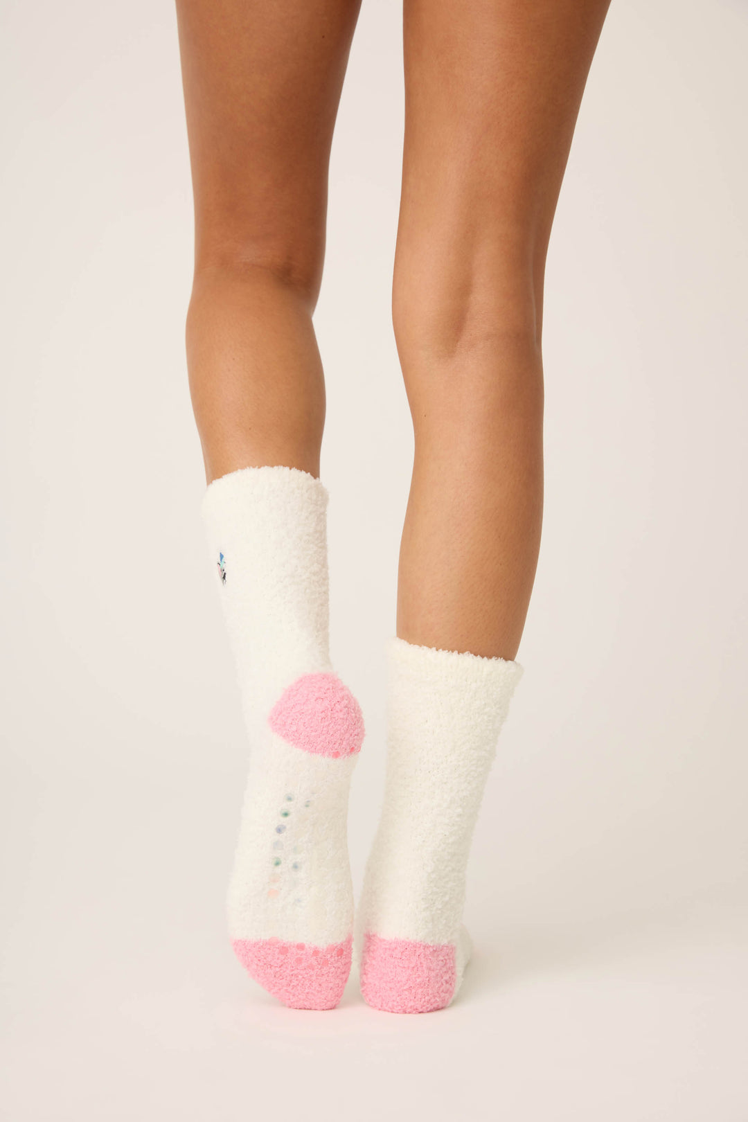 A person wears P.J. Salvage Doggone Cold Fuzzy Dog Crew Socks, featuring white fuzzy fabric, light pink heels and toes, and small rubber grips on the bottom. Only their lower legs and feet are visible on a white surface.
