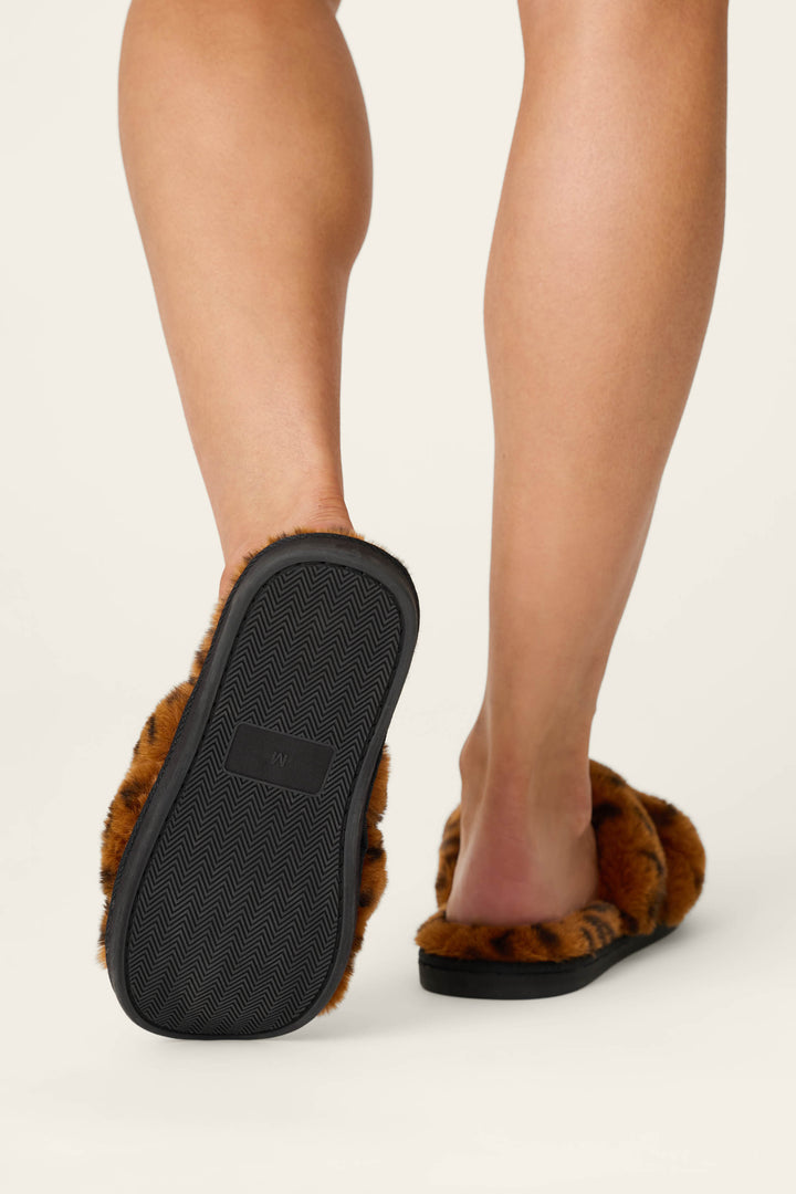 A person wearing P.J. Salvage Faux Fur Leopard Print Slippers, shown from behind with one foot raised to highlight the textured black sole against a plain background.