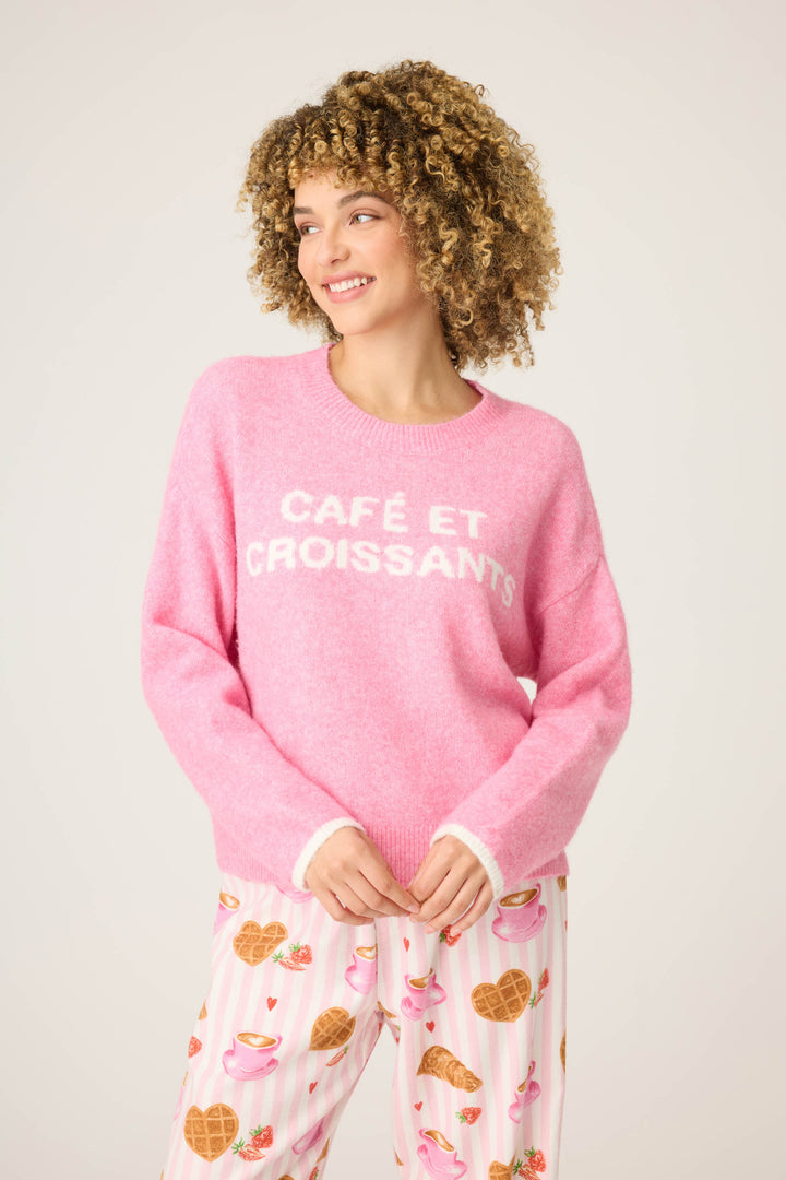 A person with curly hair wears the P.J. Salvage Cafe Et Croissants Snuggle Sweater and pajama pants with croissants and teacups, standing against a plain light background.