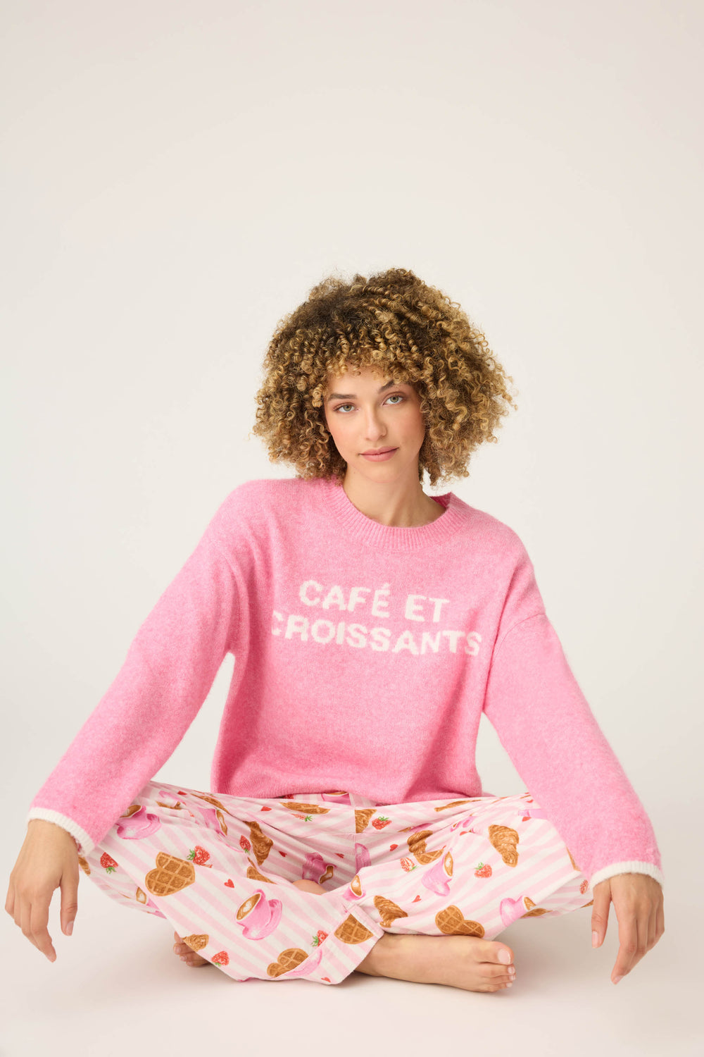 A person with curly hair sits cross-legged against a plain light background, wearing the P.J. Salvage Cafe Et Croissants Snuggle Sweater and pink pajama pants with a croissant print.