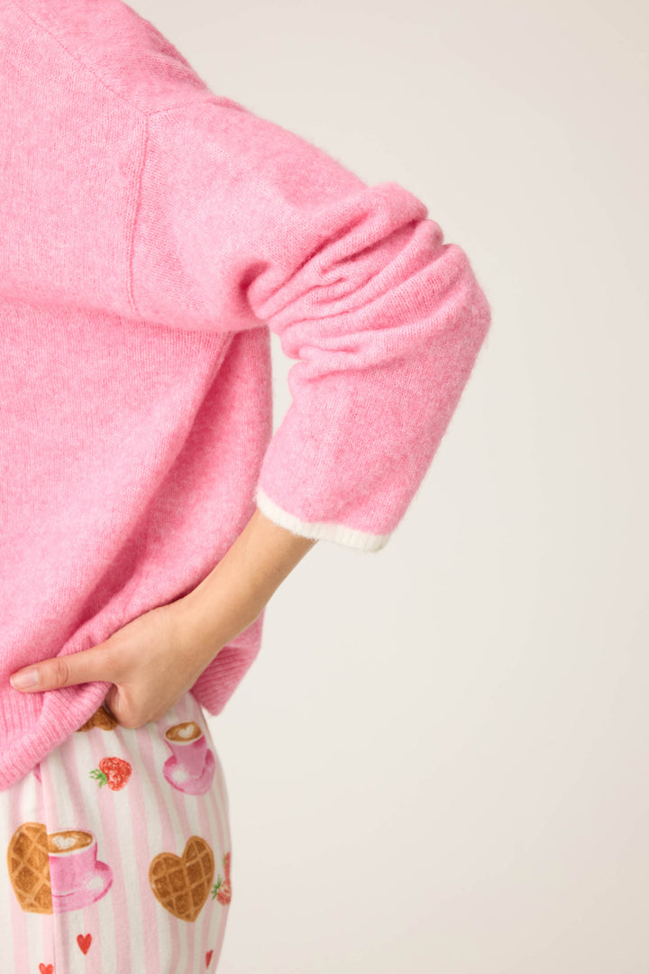 A person poses with one hand on their hip, wearing the P.J. Salvage Cafe Et Croissants Snuggle Sweater and pajama bottoms with a waffle and coffee cup pattern against a plain, light background.