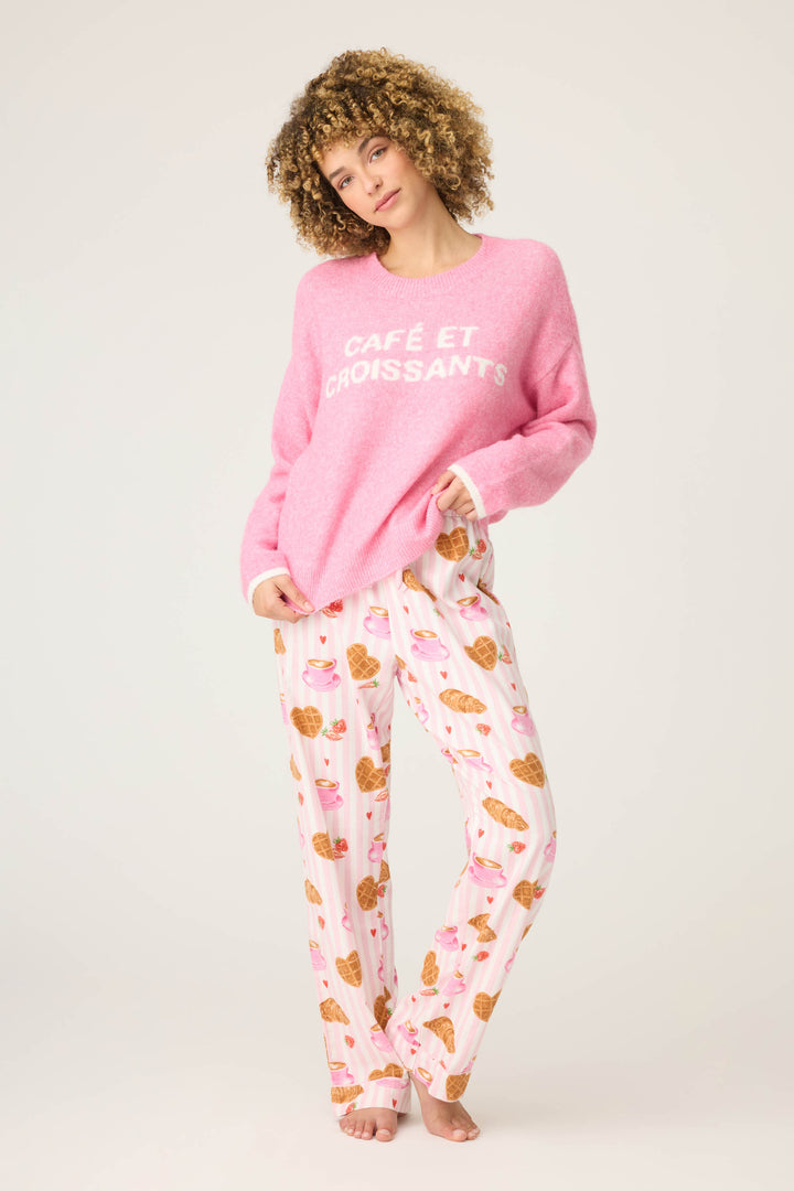 A barefoot person with curly hair stands against a plain background wearing the P.J. Salvage Cafe Et Croissants Snuggle Sweater in pink and white pajama pants with croissant and heart prints.