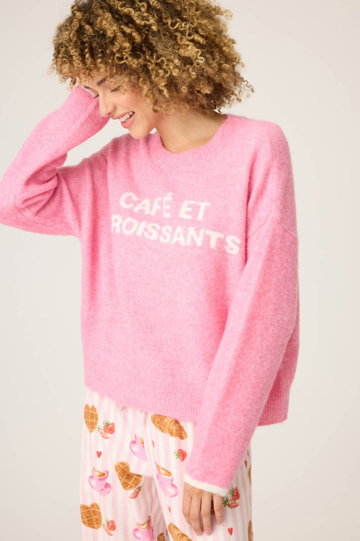 A person with curly hair wears the Cafe Et Croissants Snuggle Sweater by P.J. Salvage and patterned pajama pants with croissants, hearts, and coffee cups, standing against a light background.