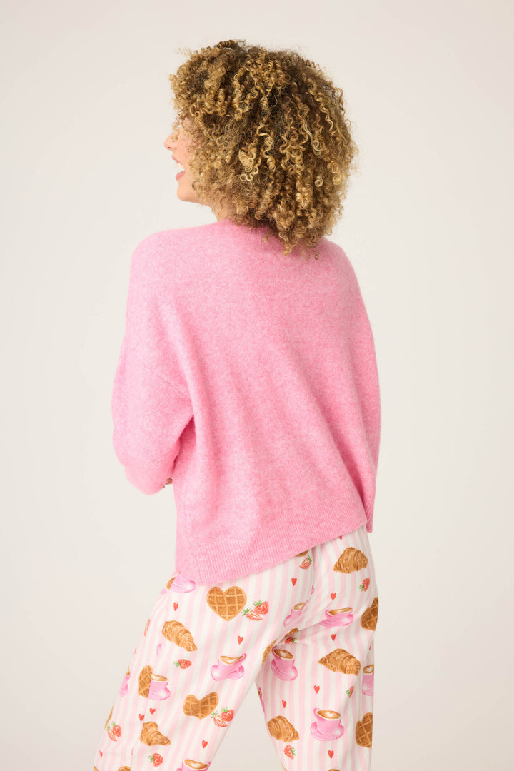 A person with curly hair, seen from behind, wears the P.J. Salvage Cafe Et Croissants Snuggle Sweater and white pajama pants featuring croissants, hearts, and teacups on a light background.