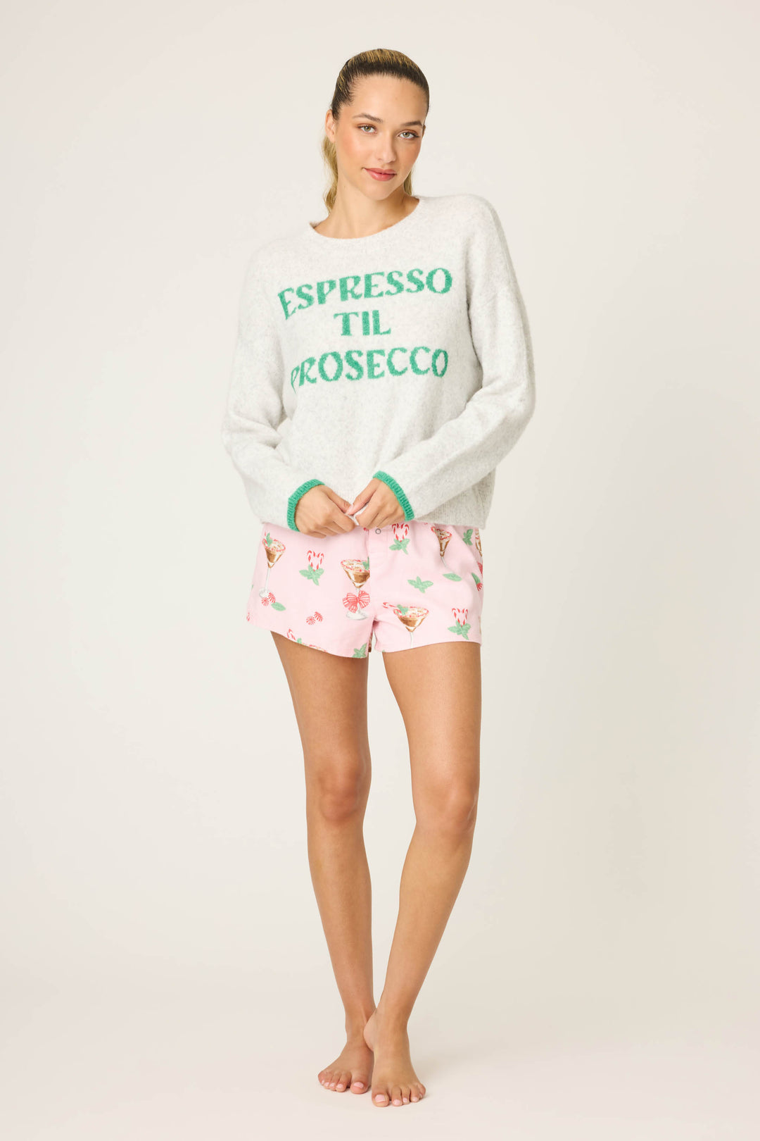A woman stands barefoot against a plain background, wearing the P.J. Salvage Espresso Til Prosecco Snuggle Sweater and light pink floral cocktail glass print shorts.