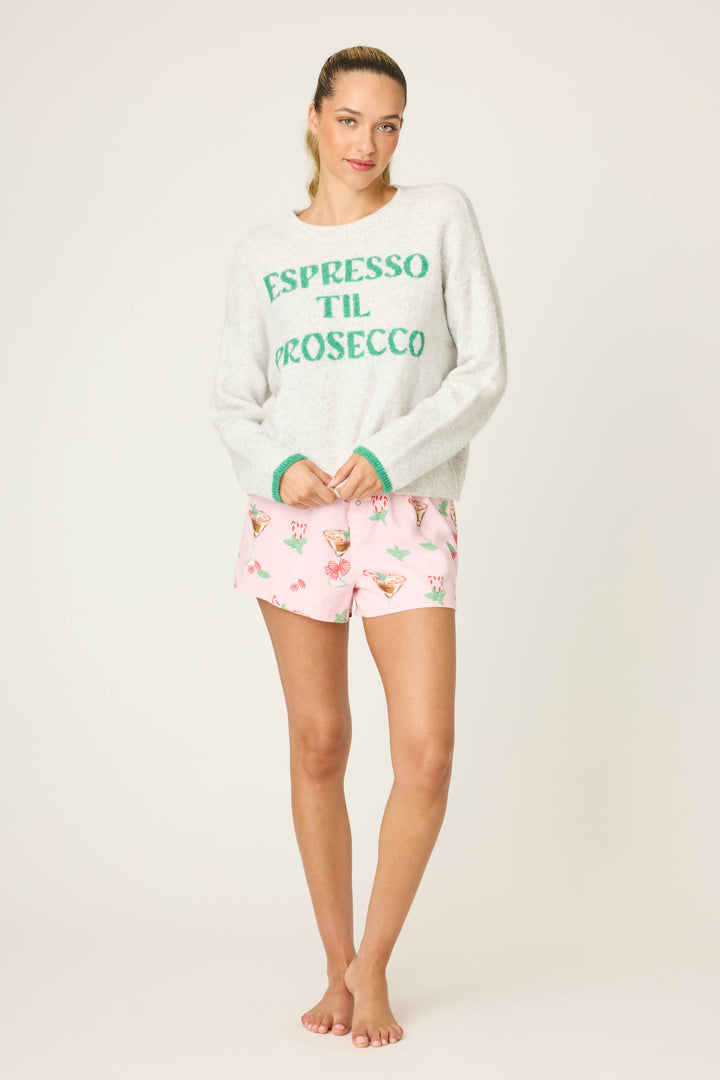 A woman stands barefoot against a plain background, wearing the P.J. Salvage Espresso Til Prosecco Snuggle Sweater and light pink floral cocktail glass print shorts.