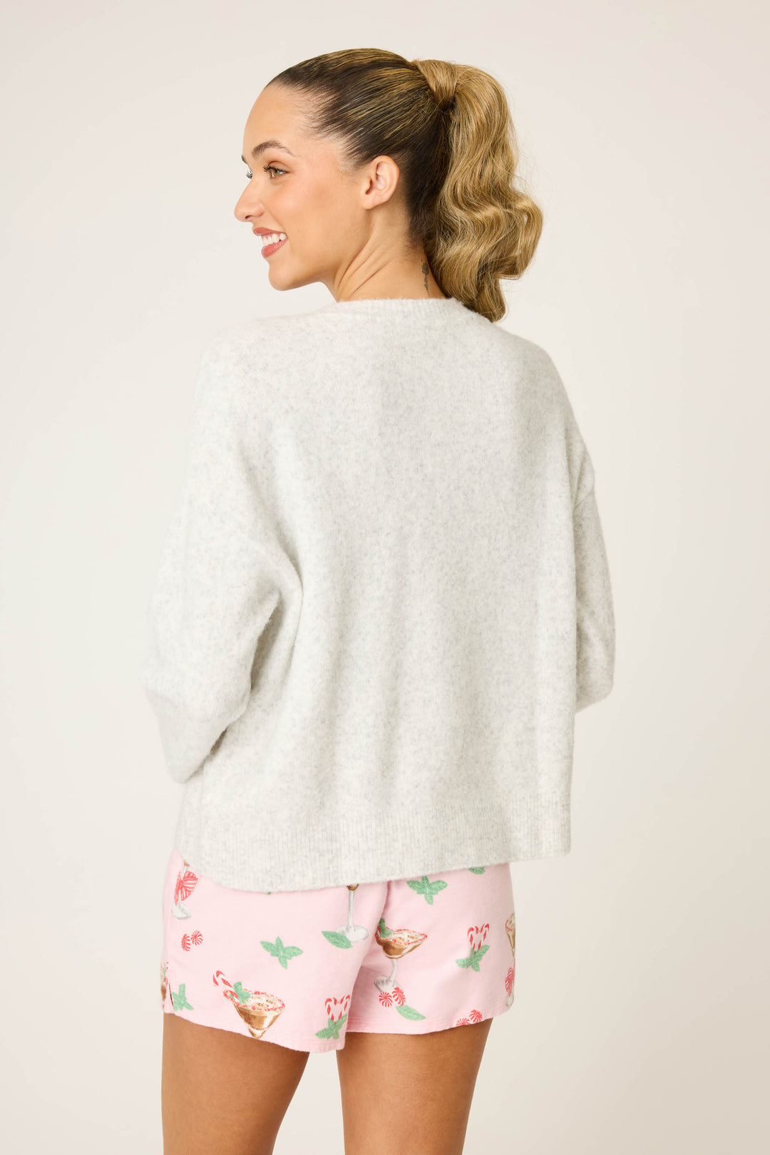 A woman with a ponytail faces away from the camera, wearing the P.J. Salvage Espresso Til Prosecco Snuggle Sweater in light gray and colorful pink shorts. The background is plain and light-colored.