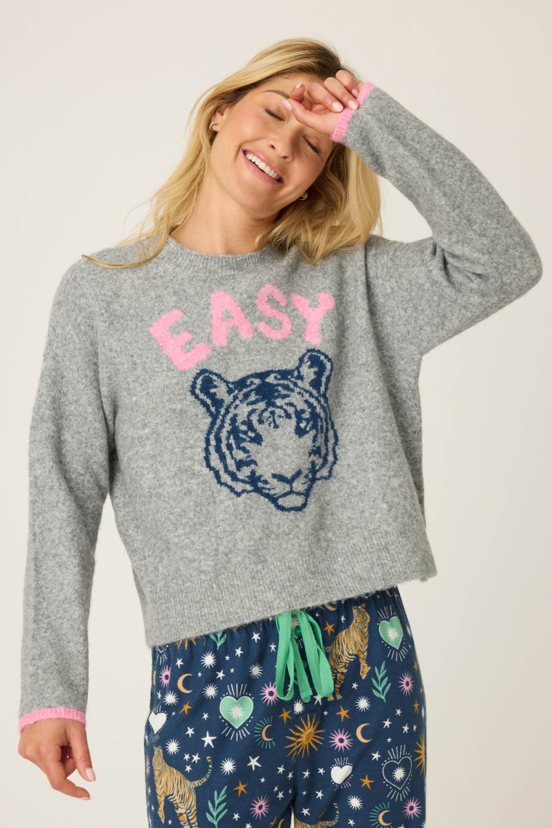 A person wears the P.J. Salvage Snuggle Sweater SLEIGH CREW LONG SLEEVE FLANNELS with a playful print, smiling and raising one hand to their forehead, paired with patterned pants featuring tigers, stars, and moons.