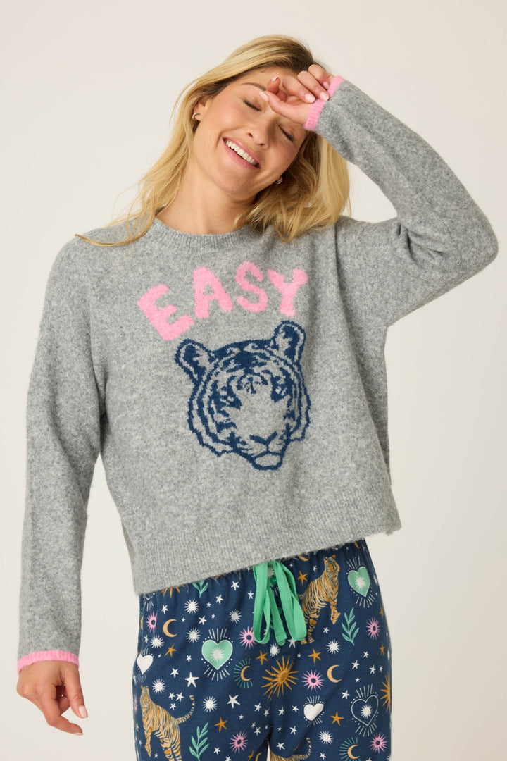 A person wears the P.J. Salvage Snuggle Sweater SLEIGH CREW LONG SLEEVE FLANNELS with a playful print, smiling and raising one hand to their forehead, paired with patterned pants featuring tigers, stars, and moons.