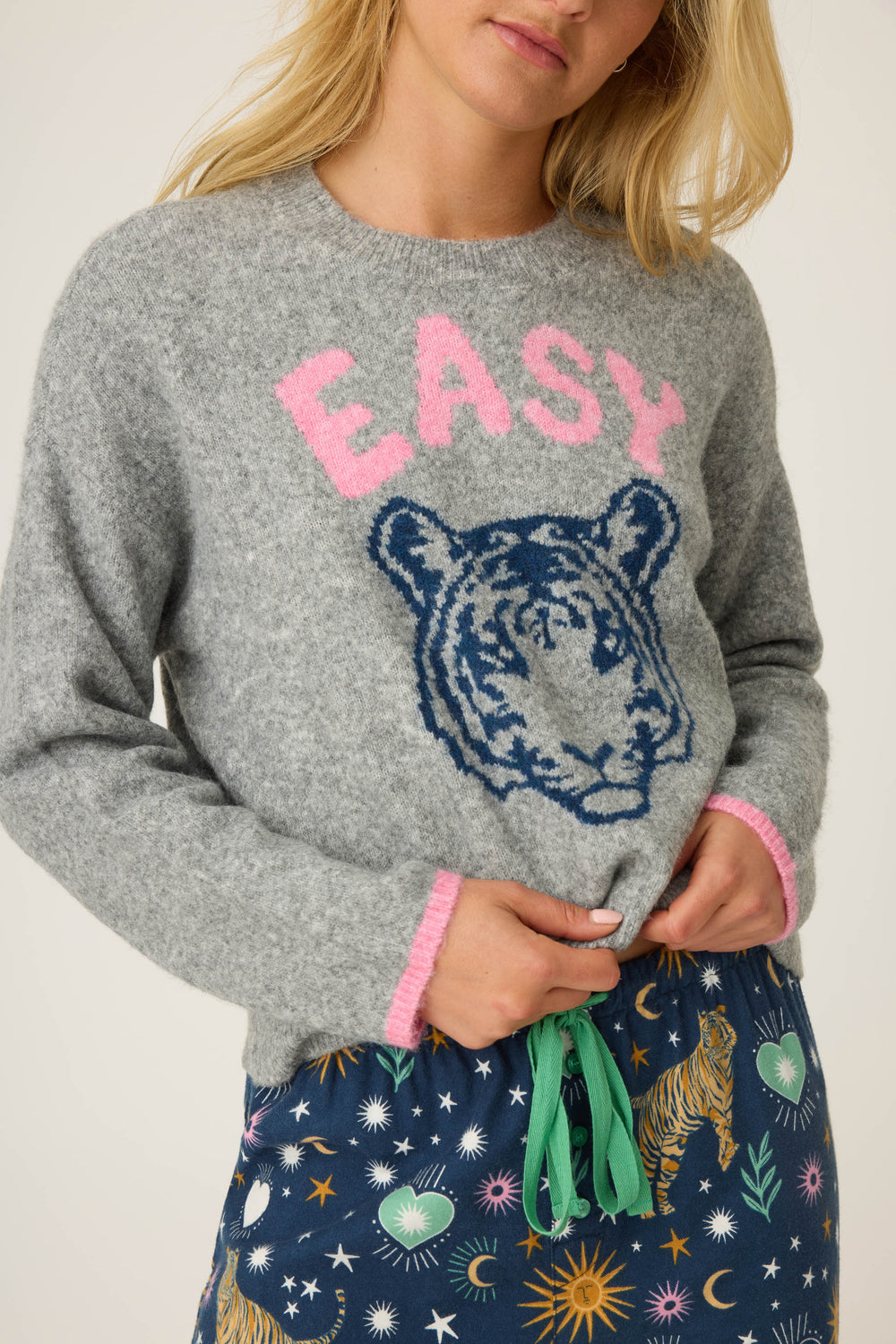 A person wears the P.J. Salvage Snuggle Sweater SLEIGH CREW LONG SLEEVE FLANNELS, a gray sweater with pink trim and EASY above a blue tiger graphic, paired with navy pants featuring green tie and colorful tigers, stars, and hearts.