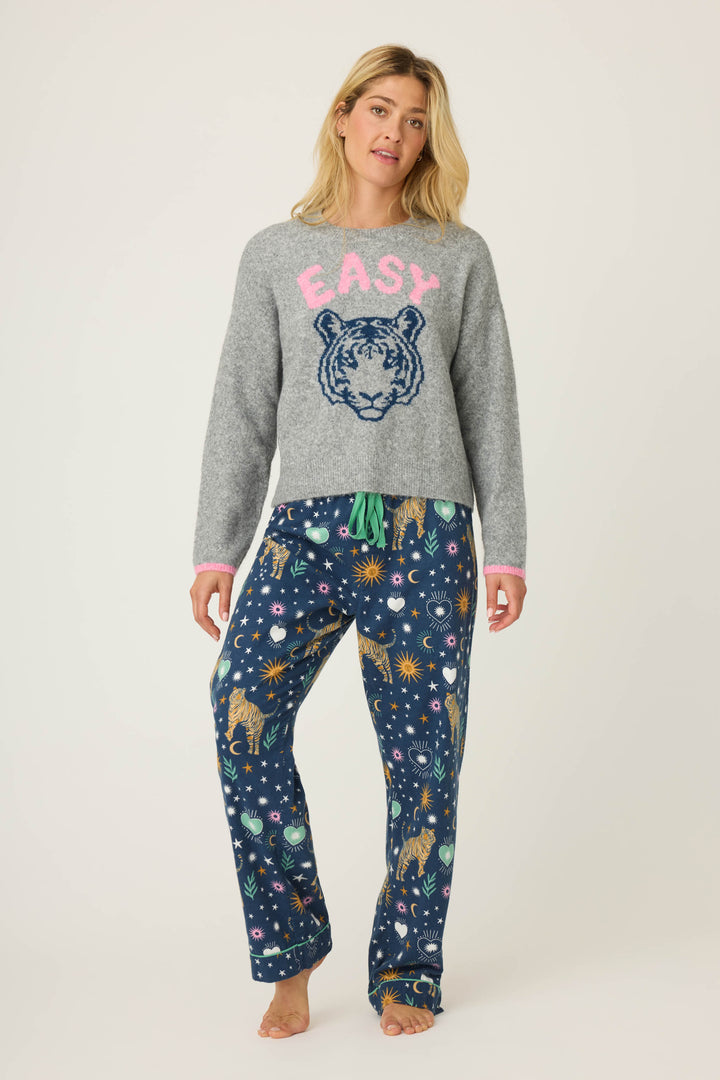 A person stands barefoot against a plain background wearing the P.J. Salvage Snuggle Sweater SLEIGH CREW LONG SLEEVE FLANNELS, a gray sweater with EASY and tiger graphic, paired with blue pajama pants featuring tigers, stars, and celestial designs.
