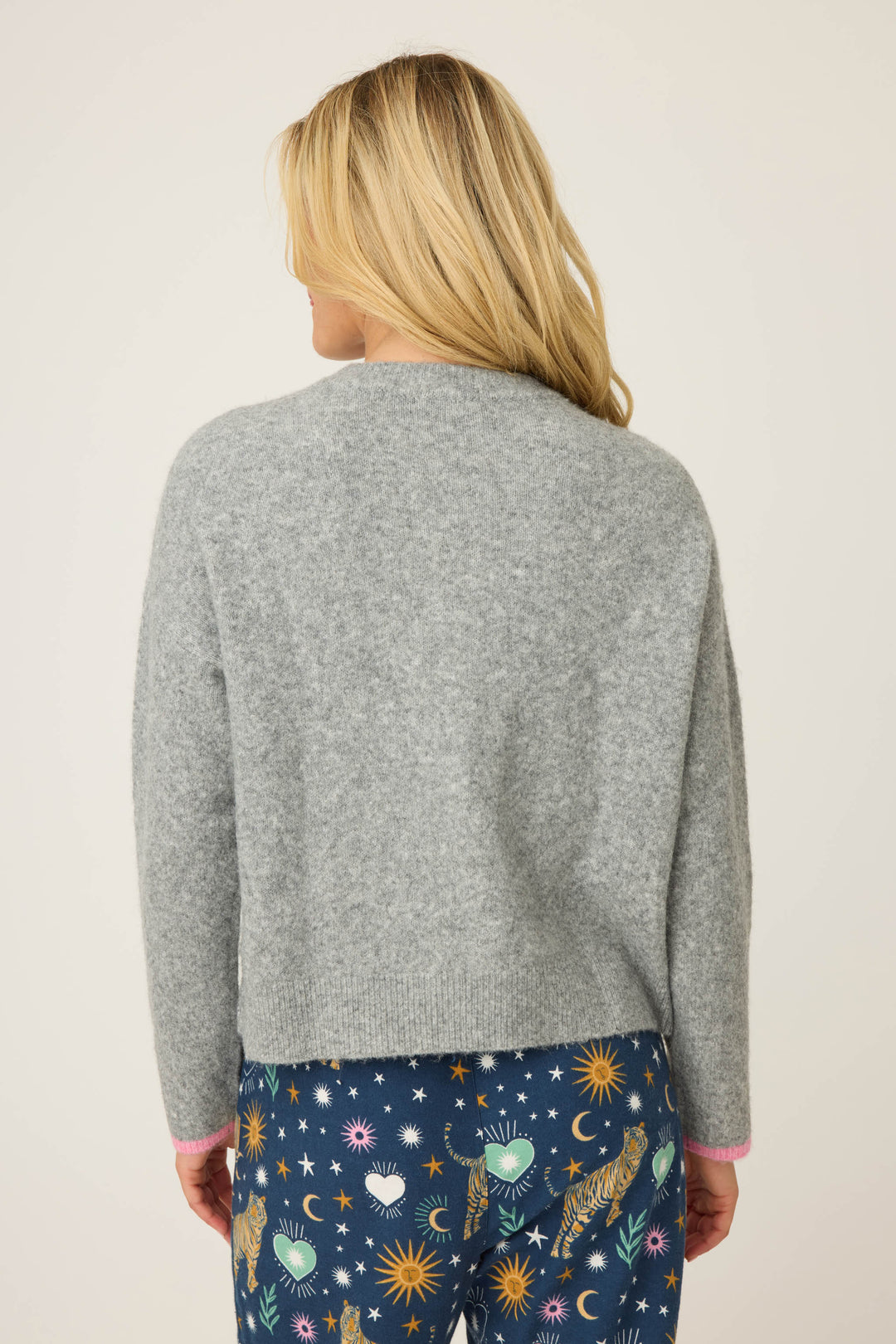 A person with long blonde hair stands with their back to the camera, wearing a P.J. Salvage Snuggle Sweater SLEIGH CREW LONG SLEEVE FLANNELS and blue pajama pants decorated with tigers, suns, moons, hearts, and stars.