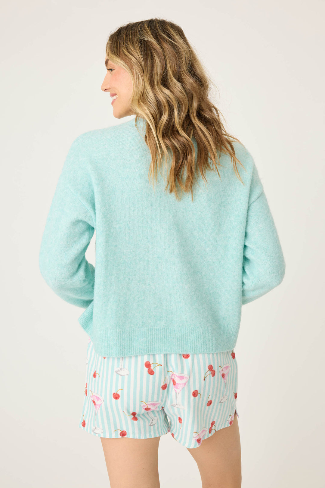 A person faces away, wearing P.J. Salvage’s Ma Cherie Snuggle Sweater in light blue with shorts featuring pink cocktails and cherries; the background is plain and light-colored.