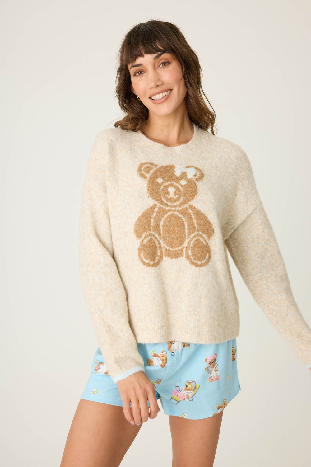 A woman with medium-length brown hair smiles, wearing the P.J. Salvage Teddy Bear Graphic Snuggle Sweater and light blue teddy bear gift print shorts, standing against a plain light background.