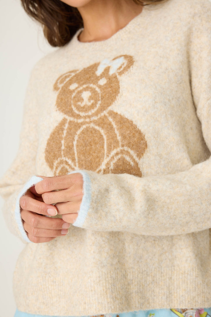 A person wearing the P.J. Salvage Teddy Bear Graphic Snuggle Sweater, a beige fuzzy top with a large teddy bear design on the front, stands with hands loosely clasped; only their torso and part of their face are visible.