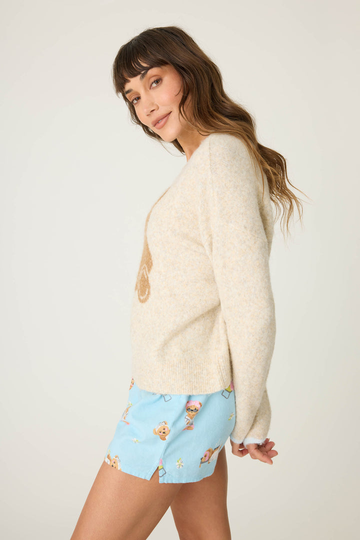 A woman with long brown hair and bangs wears the P.J. Salvage Teddy Bear Graphic Snuggle Sweater, posing sideways in a neutral expression against a plain light background. She pairs it with playful printed light blue shorts.