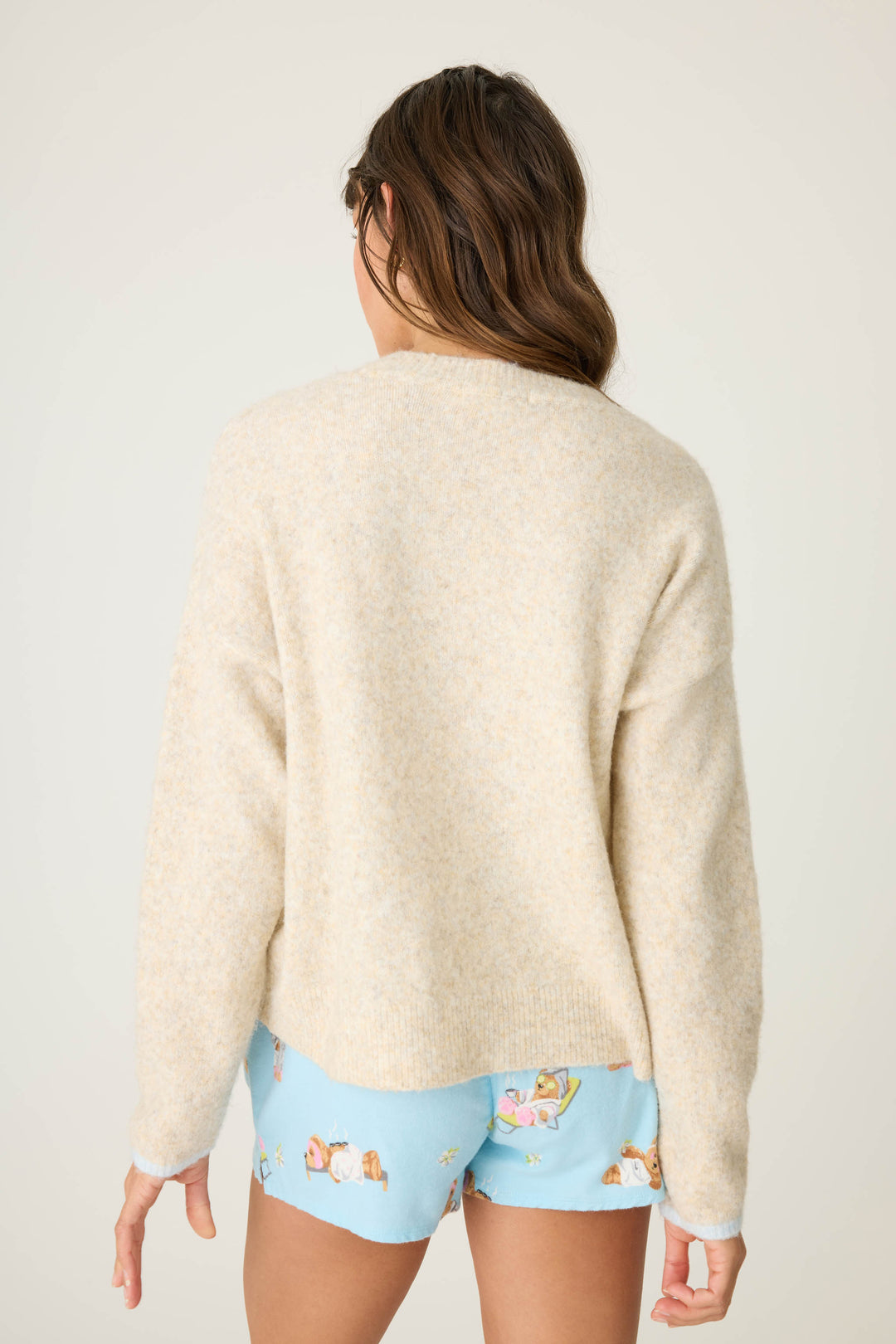 A woman with loose brown hair faces away from the camera, wearing the P.J. Salvage Teddy Bear Graphic Snuggle Sweater, cream-colored, paired with blue cartoon-print shorts against a plain, light background.