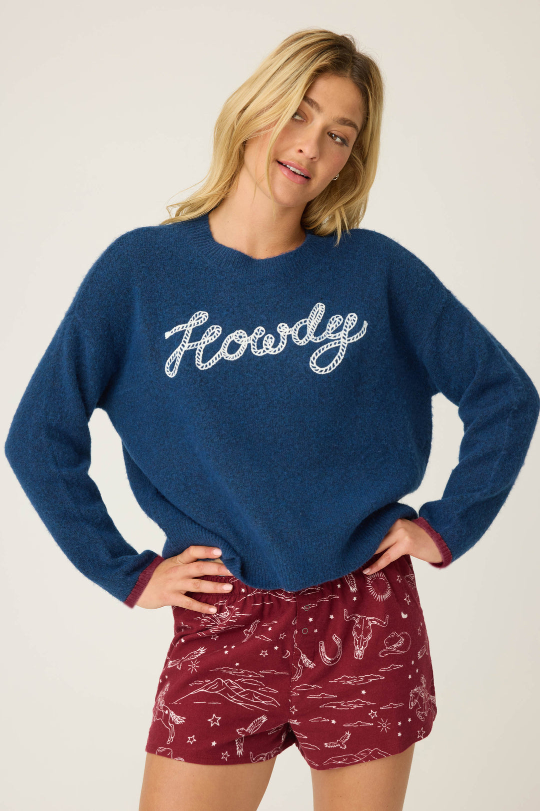 A woman with blonde hair and a neutral expression wears the P.J. Salvage Snuggle Sweater SLEIGH CREW LONG SLEEVE FLANNELS in blue and maroon shorts with a white western print, standing with her hands on her hips.
