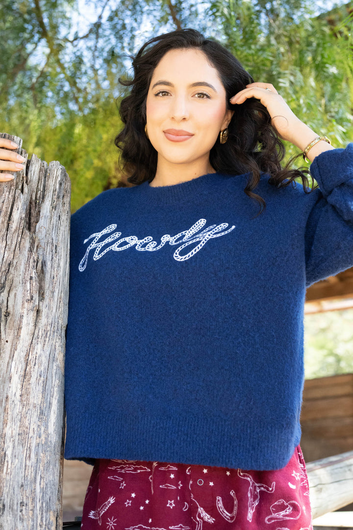 A woman with long curly hair stands outdoors by a wooden post, wearing the P.J. Salvage Snuggle Sweater SLEIGH CREW LONG SLEEVE FLANNELS. Green tree branches and a wooden structure are visible in the background.