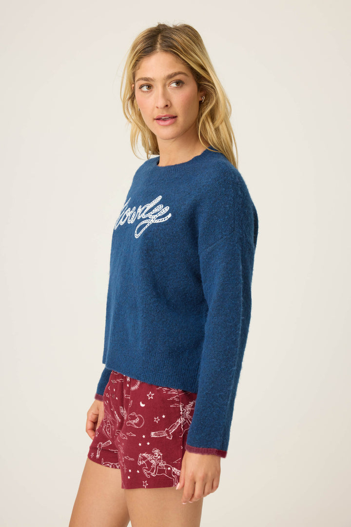 A woman with long blonde hair wears the P.J. Salvage Snuggle Sweater SLEIGH CREW LONG SLEEVE FLANNELS and red shorts with a white constellation print, standing against a plain light background.