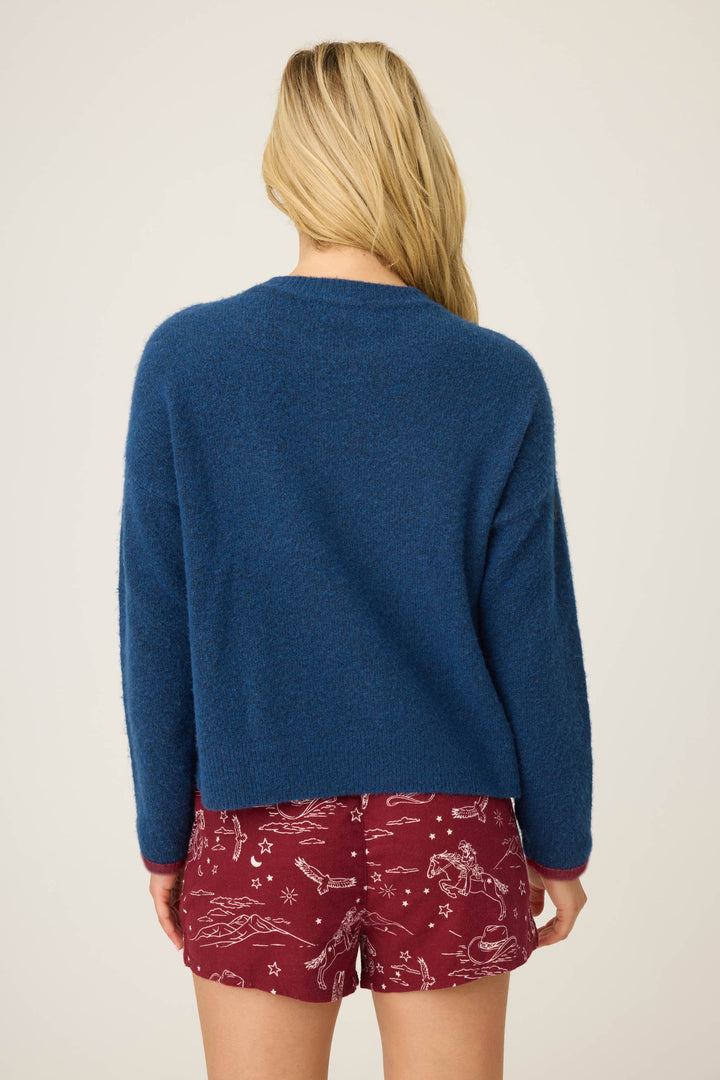 A person with long blonde hair faces away from the camera, wearing the P.J. Salvage Snuggle Sweater SLEIGH CREW LONG SLEEVE FLANNELS in blue and red shorts with a white celestial pattern, set against a plain beige background.