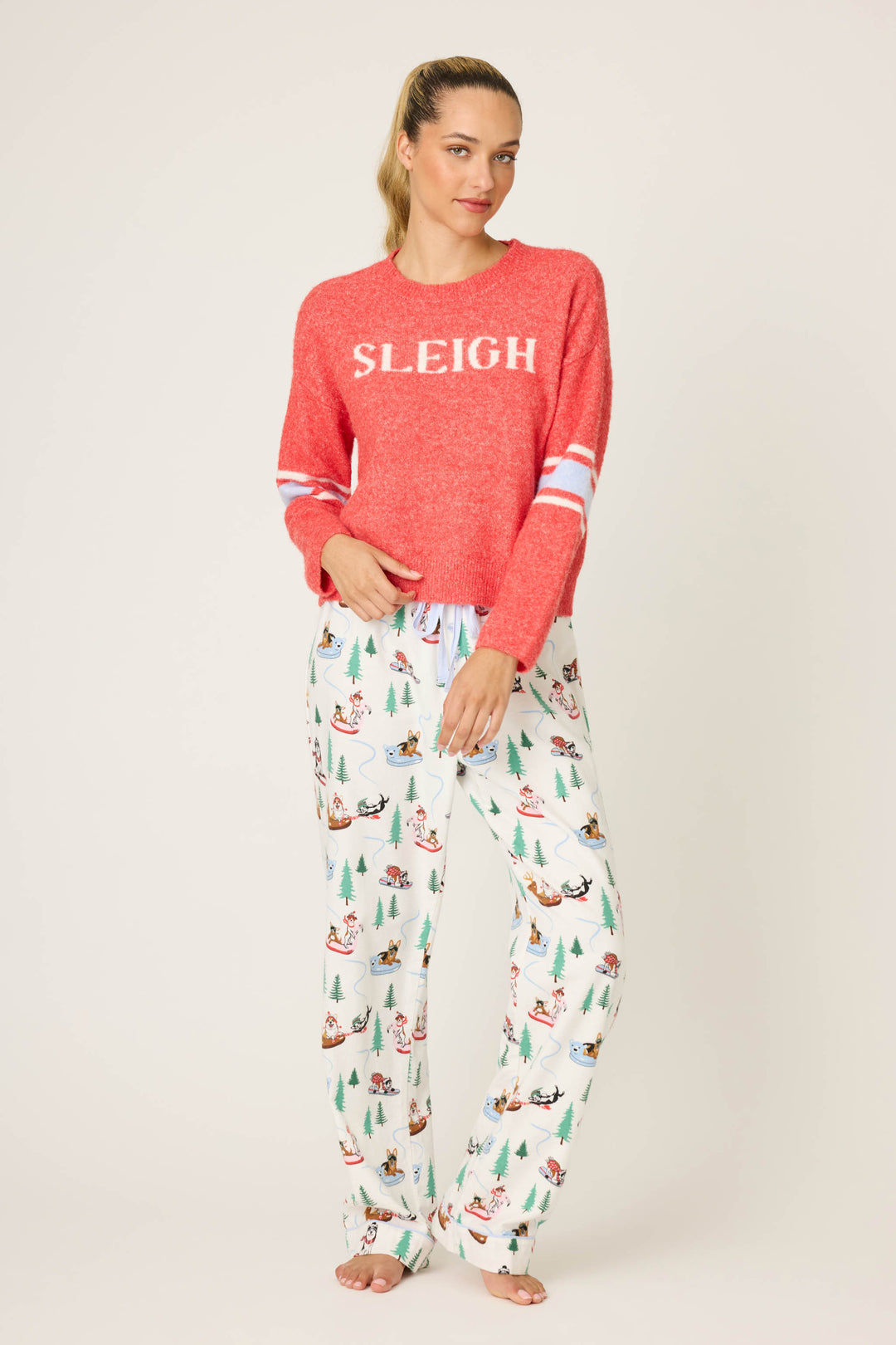 A woman with a ponytail poses against a plain background, wearing the P.J. Salvage Sleigh Snuggle Sweater in red and white holiday pajama pants. The sweater features the word "SLEIGH" on the front as she looks at the camera.