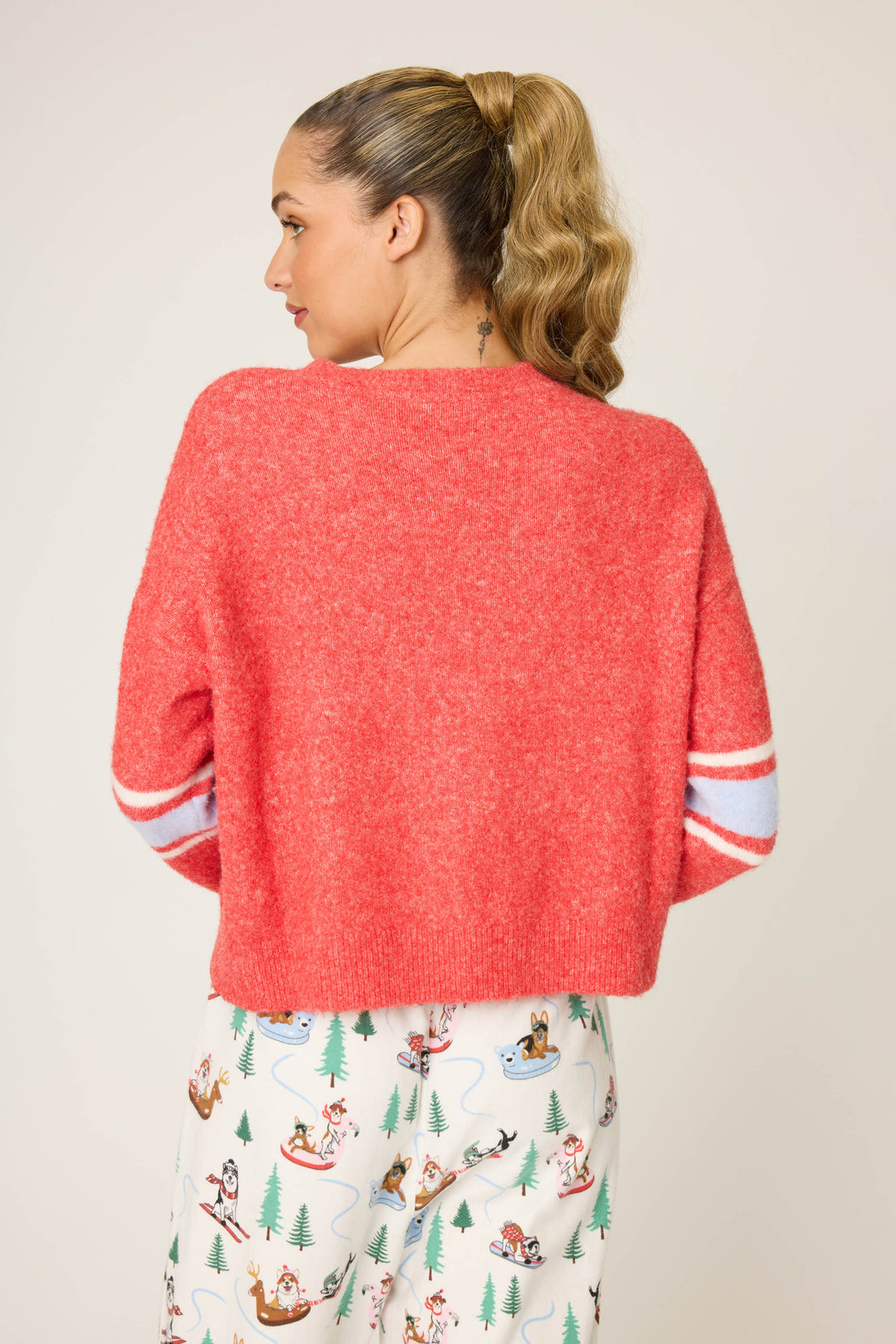 A person with a high ponytail faces away, wearing the P.J. Salvage Sleigh Snuggle Sweater—red, with white and blue sleeve stripes—paired with white pants featuring winter sports cartoon prints.