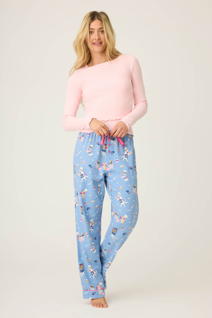 A woman with long blonde hair models P.J. Salvages WILD PANT FLANNELS—blue pajama pants with a cartoon print—paired with a light pink long-sleeve top, standing barefoot against a plain background and smiling slightly.