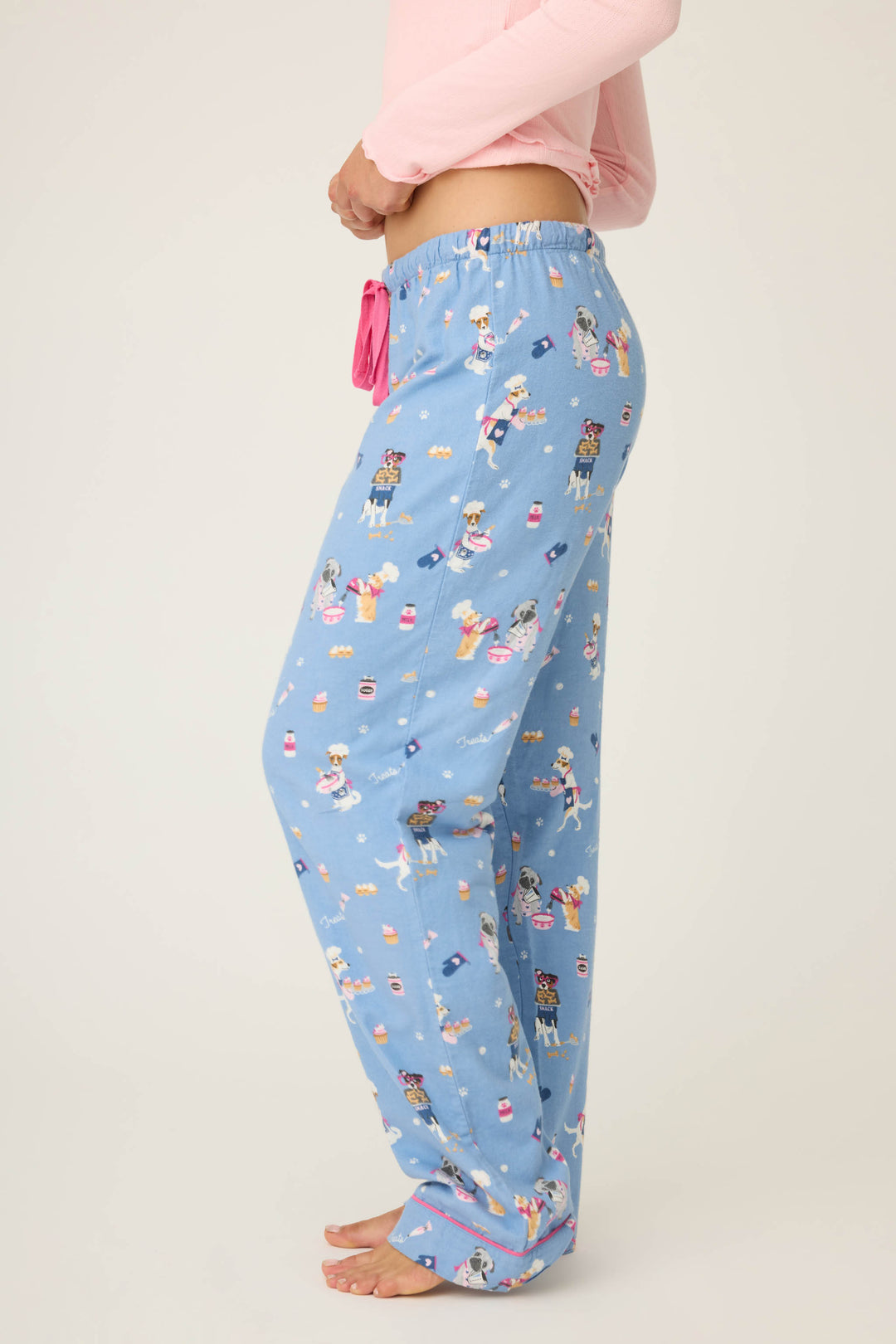 A person stands barefoot in P.J. Salvage WILD PANT FLANNELS—light blue pajama pants with playful dog print and a pink drawstring—paired with a light pink long-sleeve top, shown in side view against a plain background.