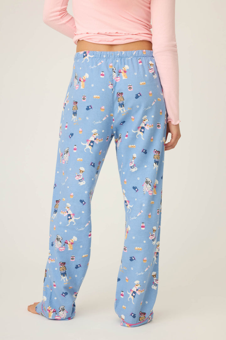 Barefoot from the waist down, a person wears P.J. Salvage WILD PANT FLANNELS—light blue pajama pants with a colorful cartoon dog and object pattern—paired with a long-sleeve pink top.