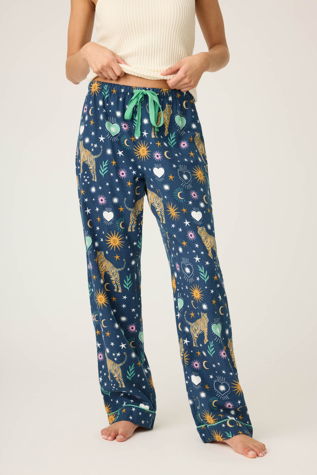 A person stands barefoot against a plain background, wearing a sleeveless ribbed cream top and P.J. Salvage WILD PANT FLANNELS—blue pajama pants with a green drawstring, adorned with stars, suns, moons, leopards, and plants.