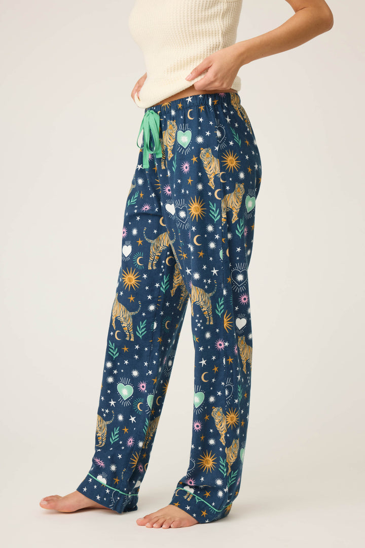 A person stands barefoot in P.J. Salvage WILD PANT FLANNELS featuring a colorful print of tigers, stars, moons, and plants with a green drawstring, paired with a cream sleeveless top.