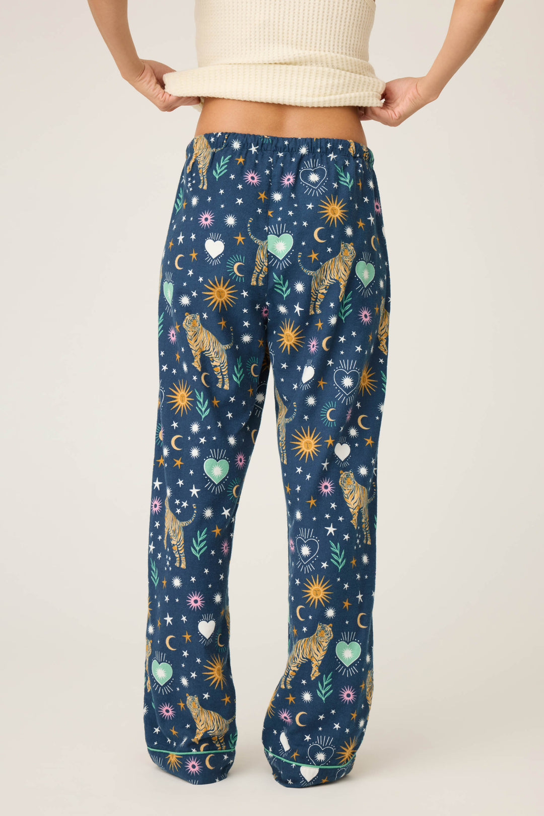 A person lifts a cream ribbed top, revealing P.J. Salvage WILD PANT FLANNELS with blue fabric and a vibrant pattern of tigers, suns, moons, stars, hearts, and flowers on a light background.