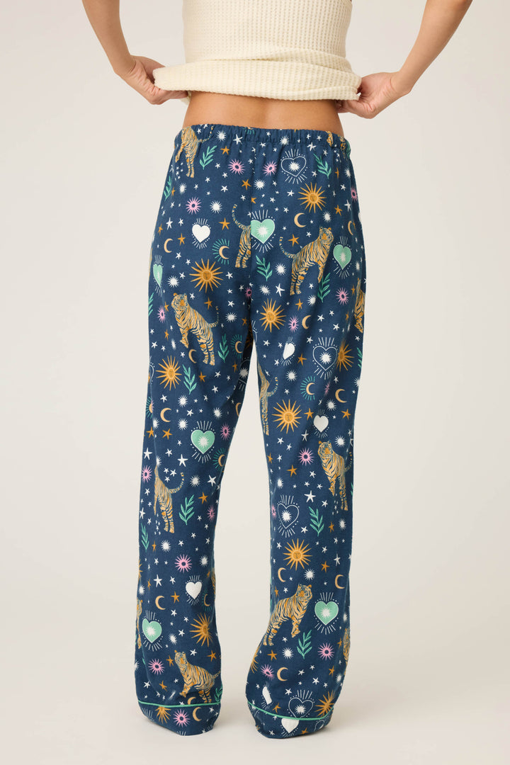 A person lifts a cream ribbed top, revealing P.J. Salvage WILD PANT FLANNELS with blue fabric and a vibrant pattern of tigers, suns, moons, stars, hearts, and flowers on a light background.
