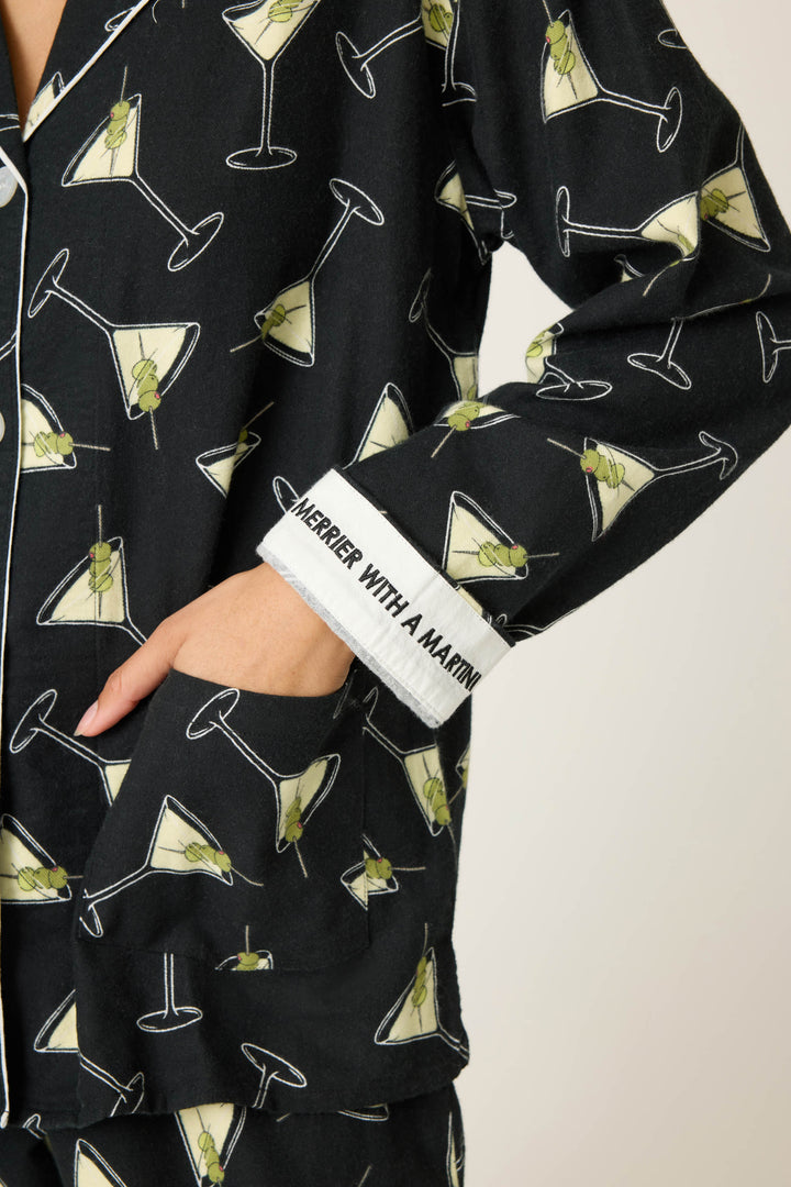 Someone wears the P.J. Salvage Merrier With A Martini Flannel PJ Set, featuring a black martini glass print. A sleeve cuff is turned up, showing embroidered "PREFER HER WITH A MARTINI." One hand rests in a pocket.