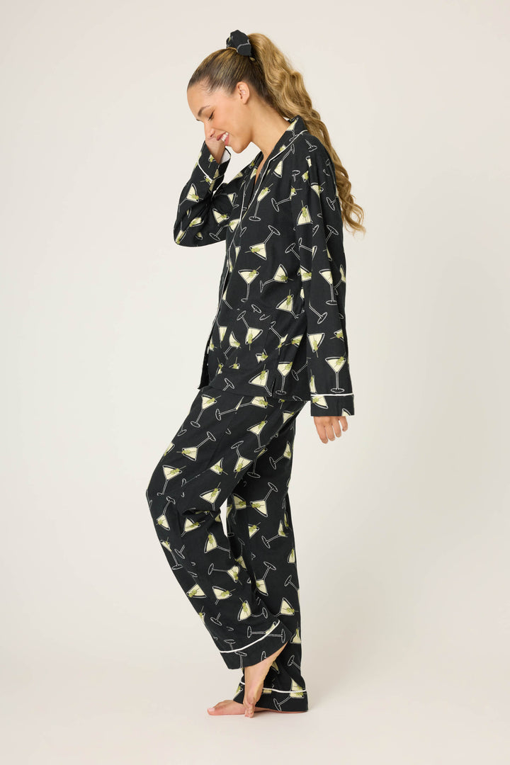 A woman with long blonde hair in a ponytail, smiling and touching her face, wears the P.J. Salvage Merrier With A Martini Flannel PJ Set featuring a martini glass print. She is barefoot against a plain background.