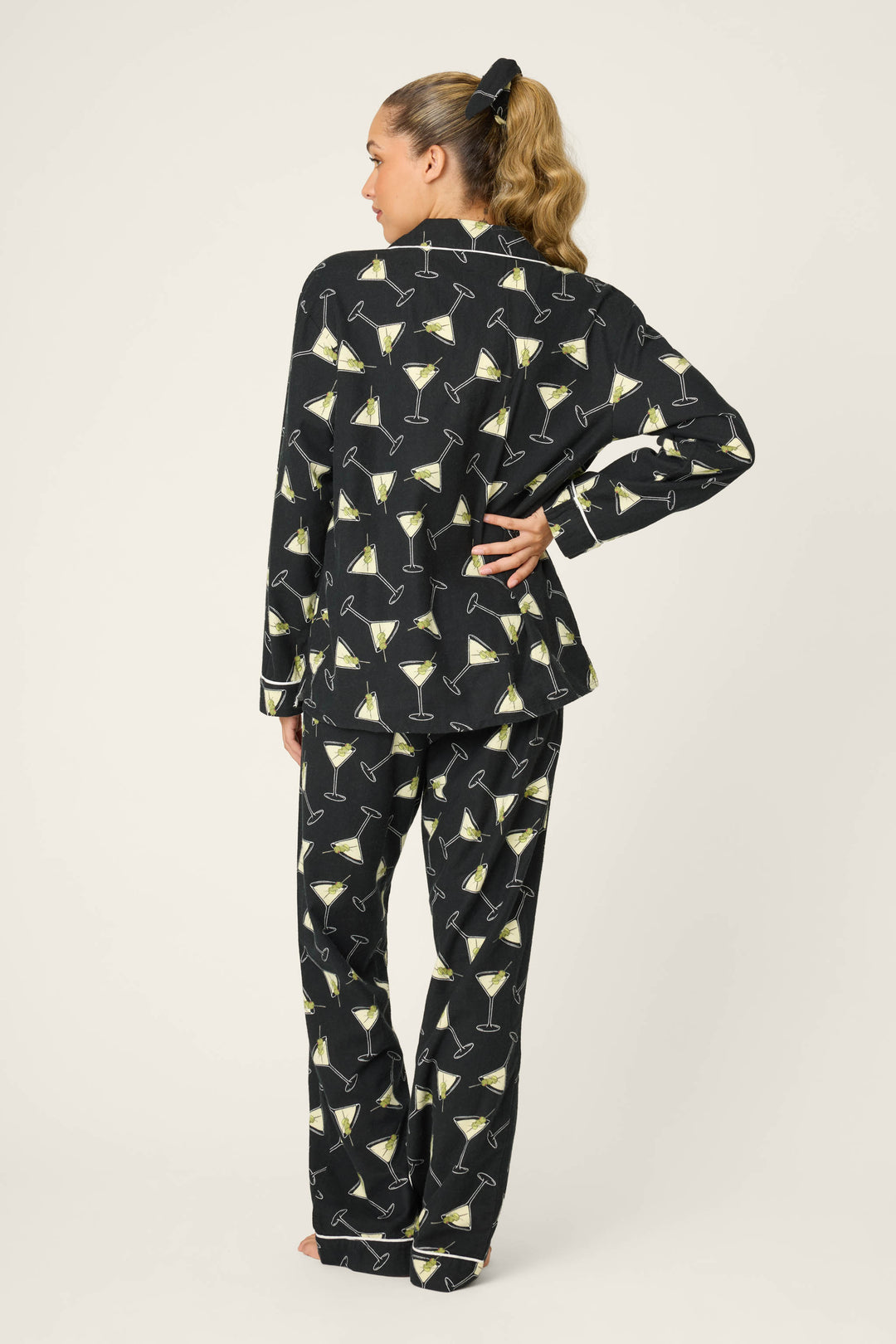 A woman faces away, wearing the P.J. Salvage "Merrier With A Martini" flannel PJ set with yellow martini glass print. Her blonde hair is tied back with a black scrunchie against a plain, light background.