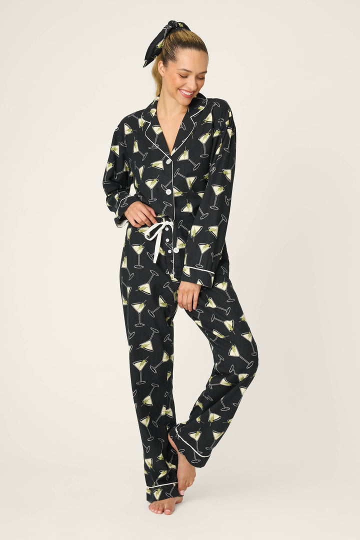 A woman smiles barefoot in the P.J. Salvage Merrier With A Martini Flannel PJ Set, featuring black pajama pants and a matching button-up top with a martini glass print. She poses with one leg bent and wears a matching hair tie.
