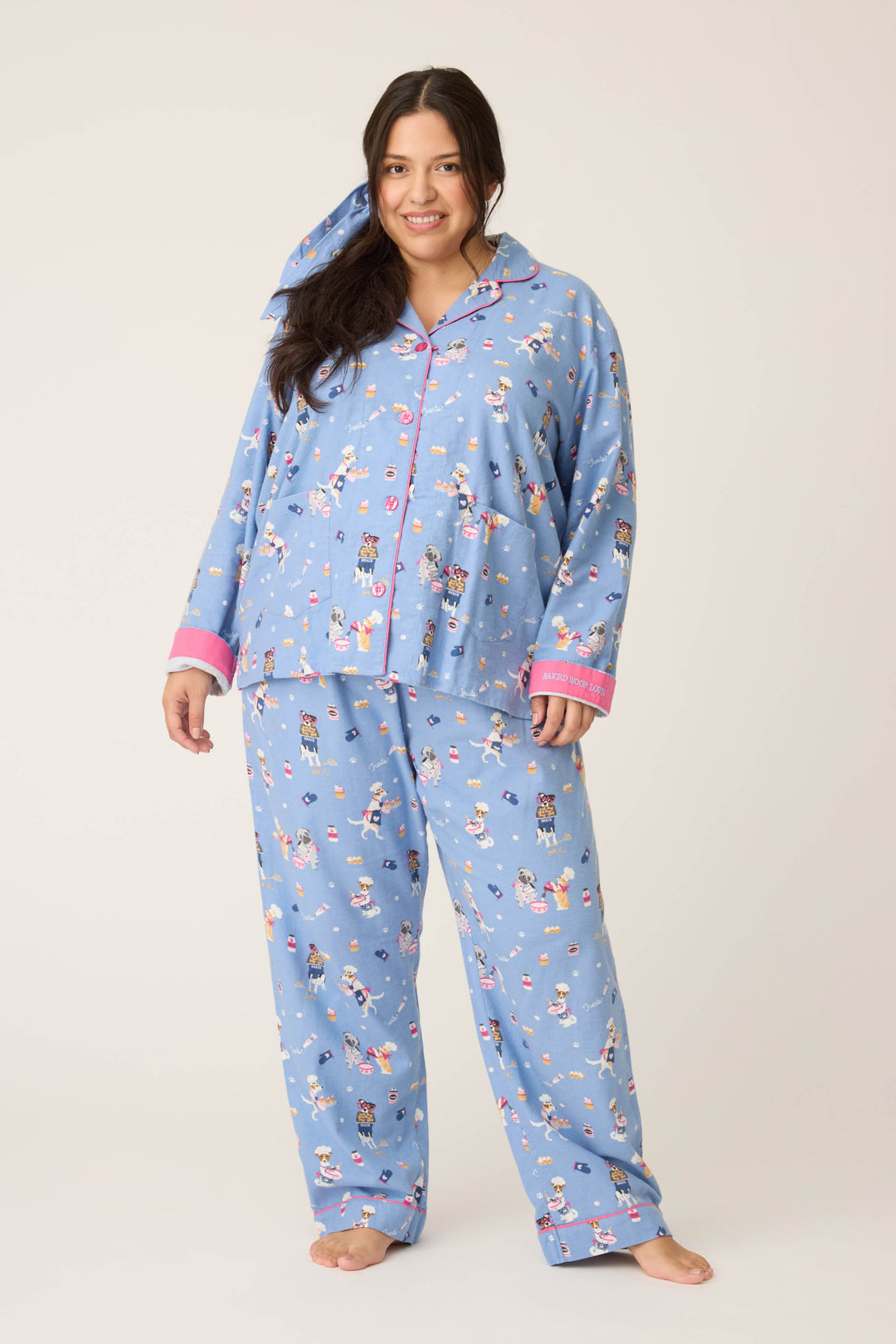 A woman with long dark hair stands barefoot, smiling, and wearing the P.J. Salvage 3-Piece Set WILD PJ SET FLANNELS PLUS—light blue pajamas featuring multicolored cartoon prints and pink-trimmed cuffs and collar—against a plain background.
