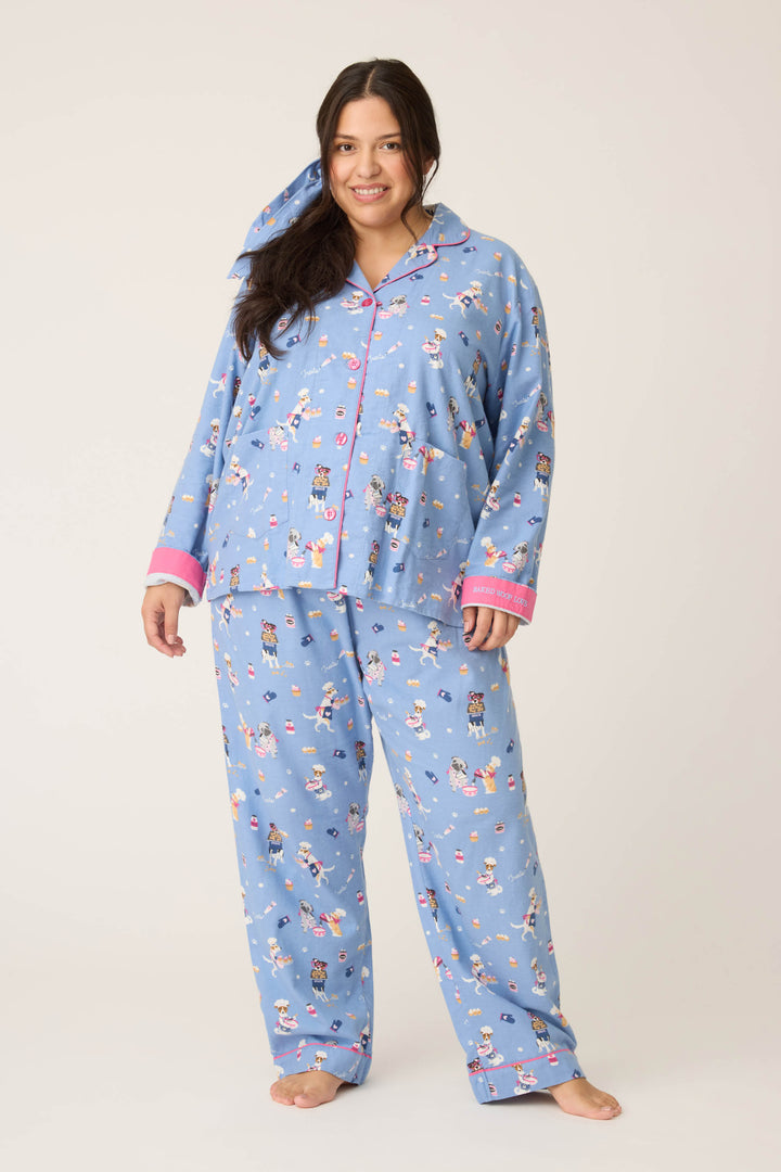 A woman with long dark hair stands barefoot, smiling, and wearing the P.J. Salvage 3-Piece Set WILD PJ SET FLANNELS PLUS—light blue pajamas featuring multicolored cartoon prints and pink-trimmed cuffs and collar—against a plain background.