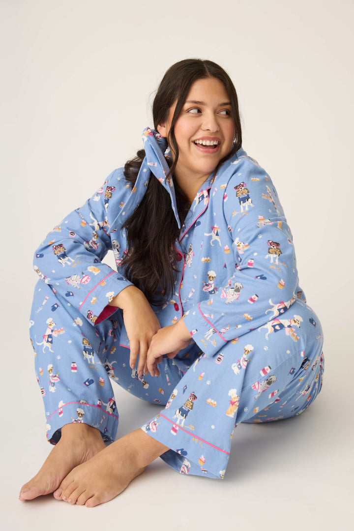 A woman with long dark hair sits barefoot on the floor, smiling and looking to the side against a plain light background. Shes wearing the P.J. Salvage 3-Piece Set WILD PJ SET FLANNELS PLUS in blue with a playful dog print.