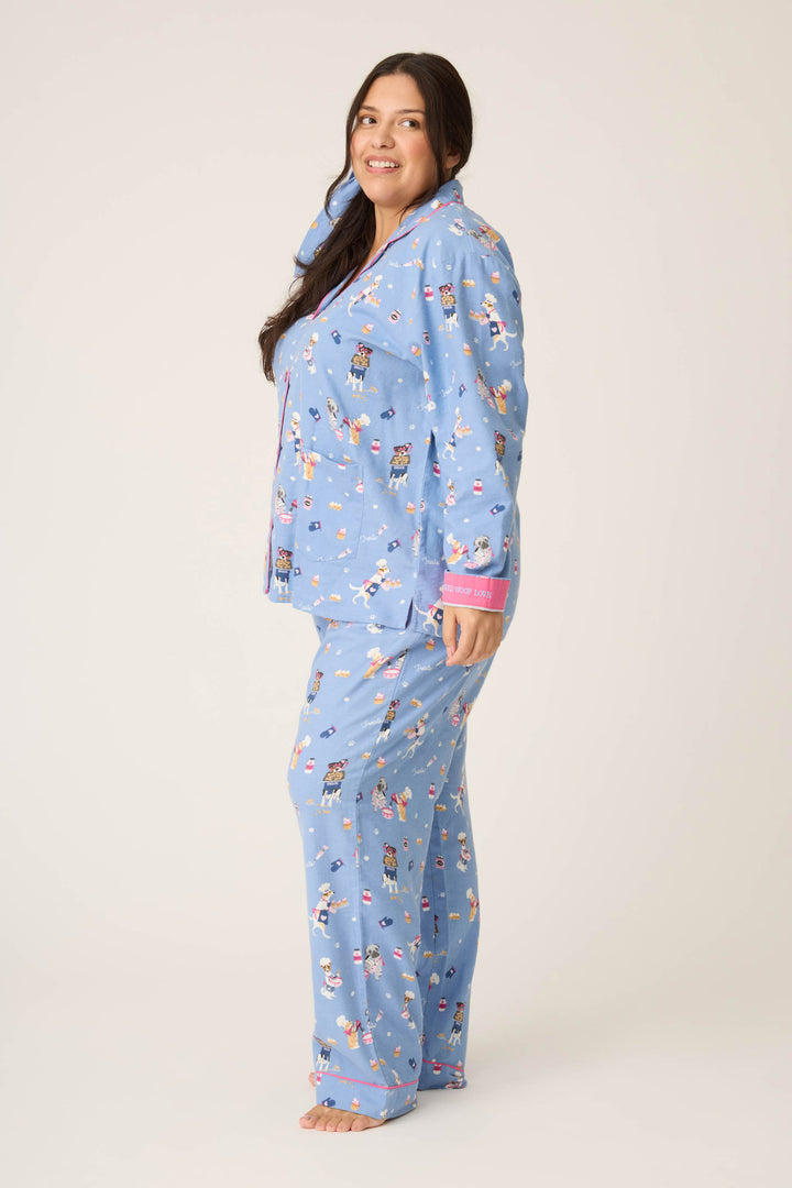 A woman with long dark hair stands barefoot against a plain background, smiling and facing slightly to the side. She wears the P.J. Salvage 3-Piece Set WILD PJ SET FLANNELS PLUS, a blue pajama set featuring a colorful dog print.
