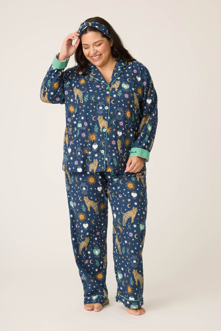 A woman smiles while looking down, barefoot in P.J. Salvage’s 3-Piece Set WILD PJ SET FLANNELS PLUS—matching navy blue pajamas and headband with celestial and tiger prints.