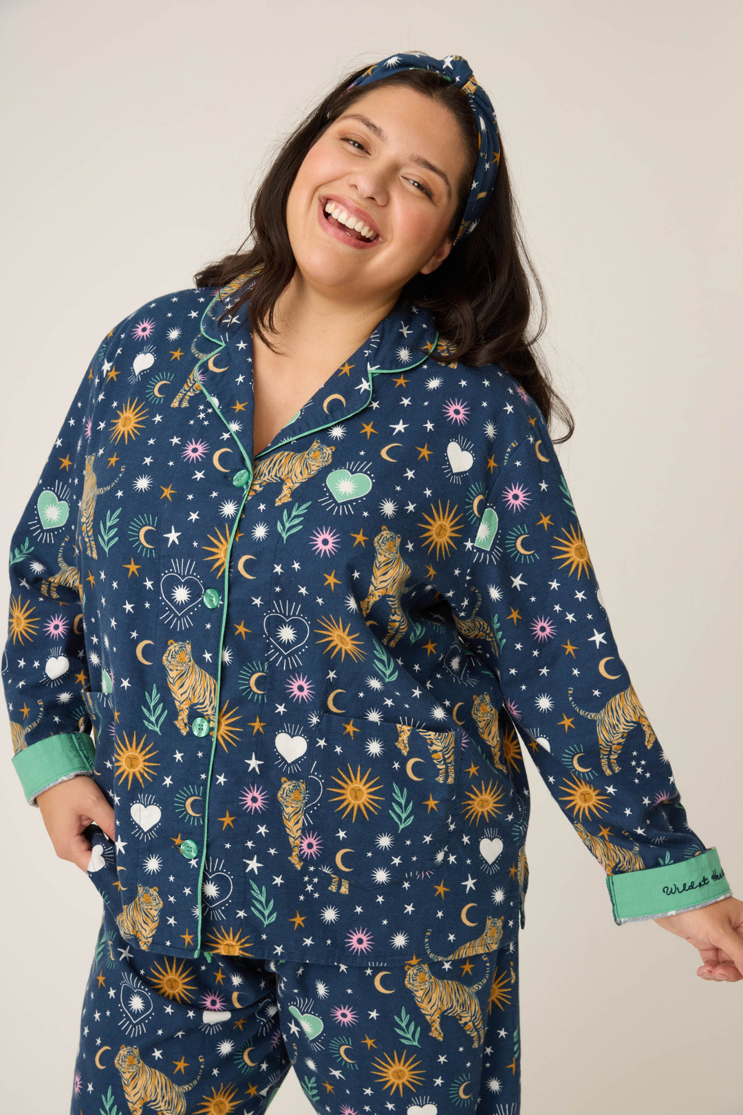 A woman smiles in the P.J. Salvage 3-Piece Set WILD PJ SET FLANNELS PLUS, featuring celestial and tiger prints. She pulls back her sleeve to show embroidered text and wears the matching headband.