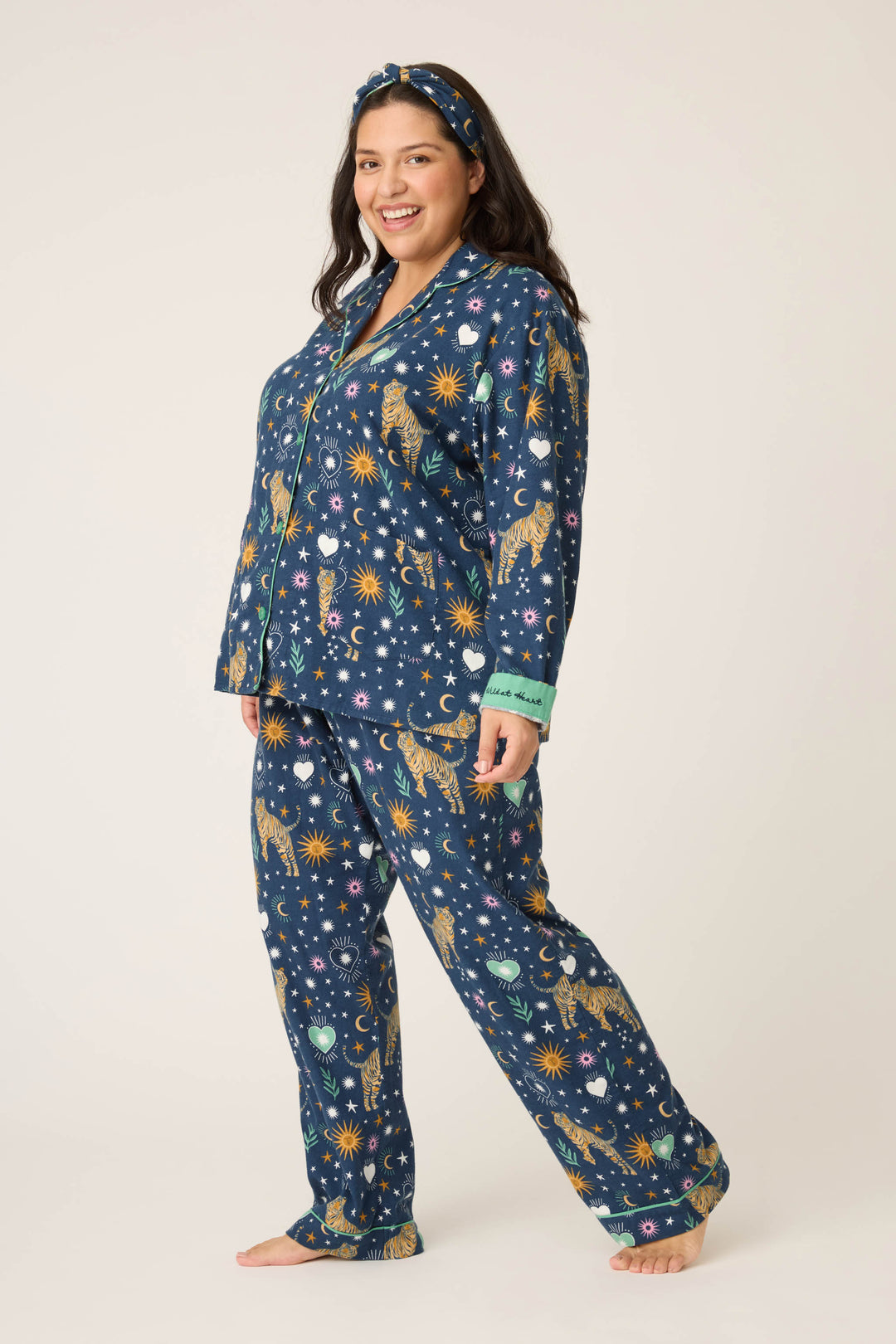 A woman smiles barefoot in a matching 3-Piece Set WILD PJ SET FLANNELS PLUS by P.J. Salvage, featuring navy blue celestial and animal prints with a coordinating headband. The background is plain and light-colored.