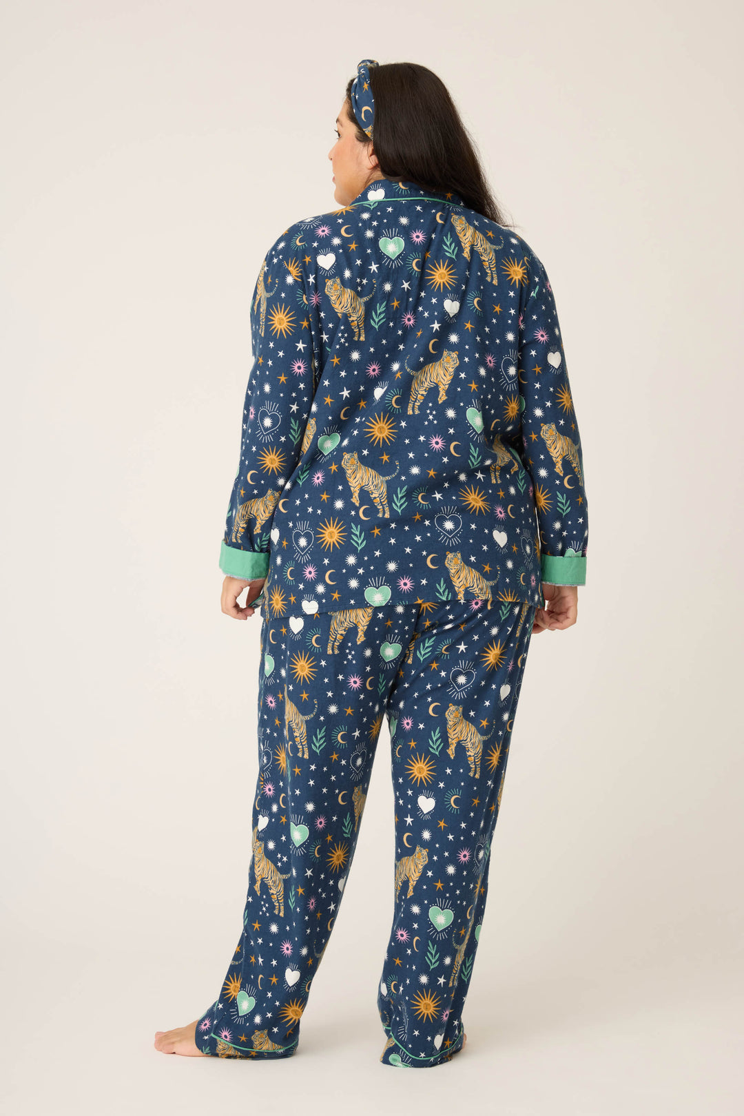 A person with long dark hair stands barefoot, wearing the P.J. Salvage 3-Piece Set WILD PJ SET FLANNELS PLUS—navy blue pajamas with celestial and big cat prints, green cuffs, and a matching headband.