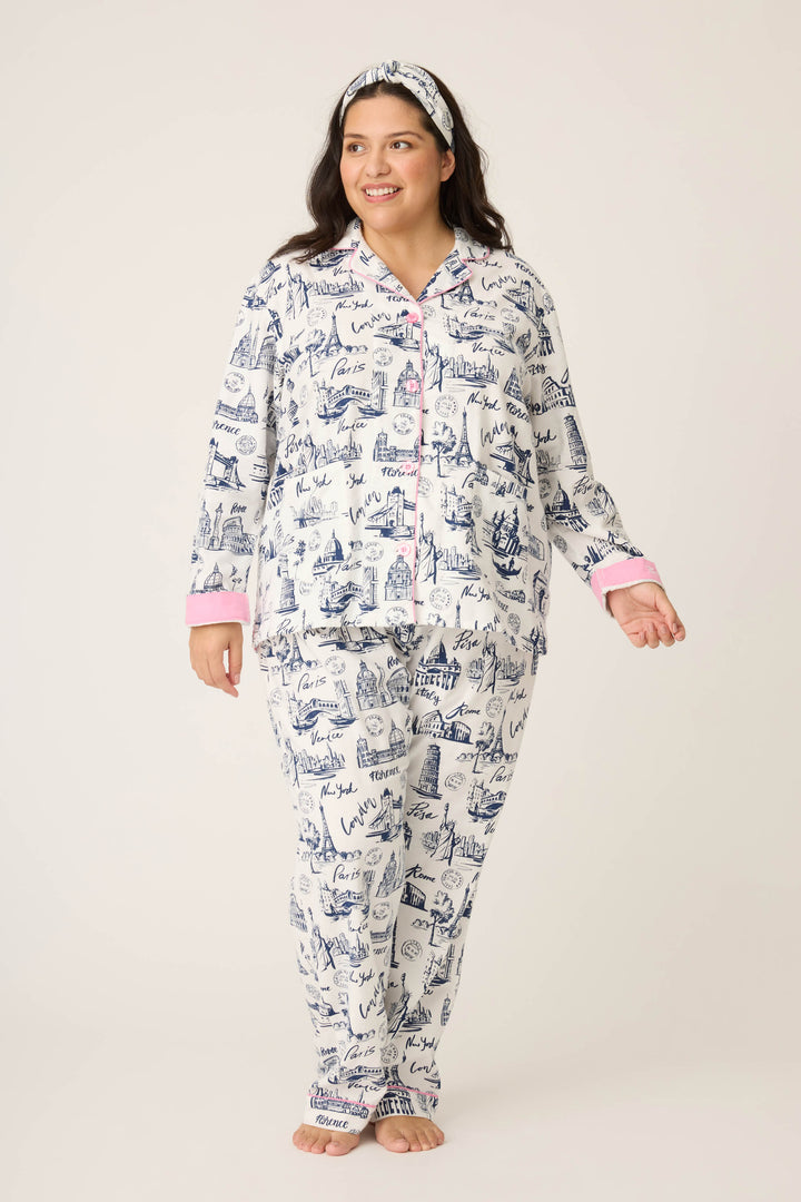 A woman stands barefoot against a plain background, smiling as she looks to the side in the P.J. Salvage 3-Piece Set WILD PJ SET FLANNELS PLUS—white pajamas with blue sketches, pink accents, and a matching headband.