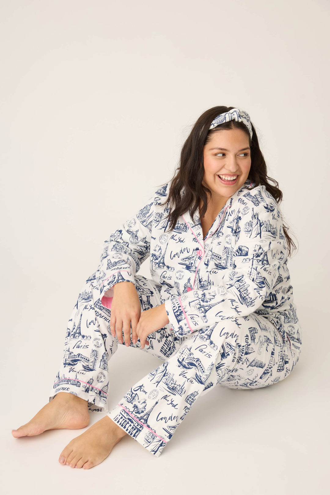 A woman with long dark hair smiles while sitting on the floor, wearing a matching headband and the P.J. Salvage 3-Piece Set WILD PJ SET FLANNELS PLUS. The background is plain and light-colored.