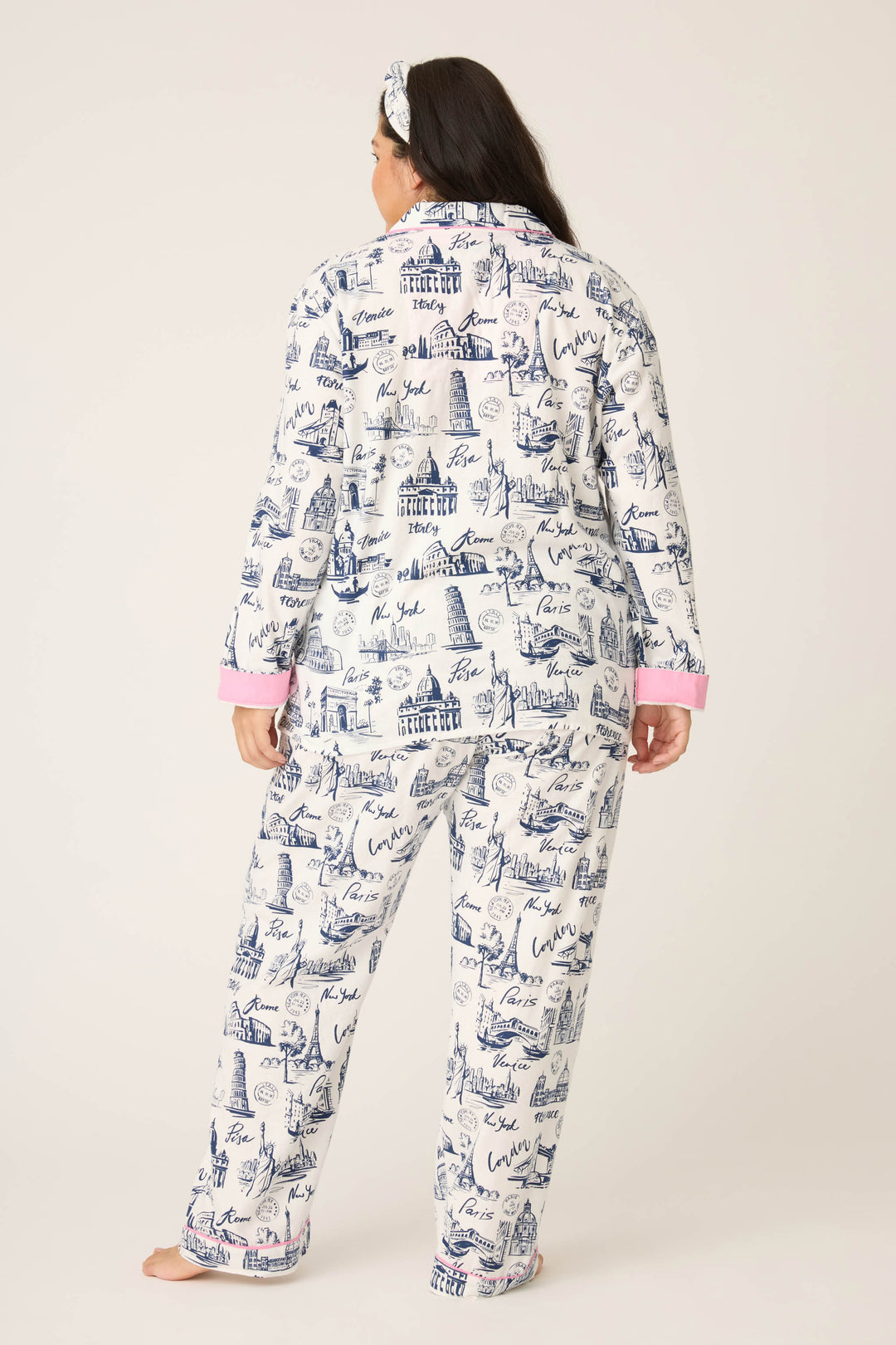 A person stands barefoot against a plain, light background wearing P.J. Salvages 3-Piece Set WILD PJ SET FLANNELS PLUS: white pajamas with a blue cityscape print, pink cuffs, long sleeves and pants, plus a matching headband.